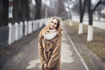 Obraz premium Blond-haired young girl cheerful joyful dressed you in a beige coat and white scarf. Autumn gray street on the background. empty road