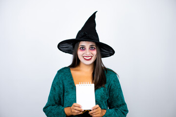 Woman wearing witch costume over isolated white background smiling and showing blank notebook in her hand