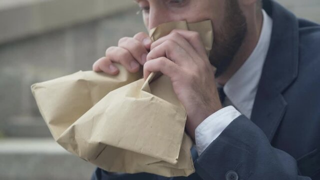 Close-up of frustrated nervous man trying to calm down by breathing into paper bag. Stressed Caucasian businessman having anxiety attack outdoors sitting on urban city stairs. Stress and overworking.