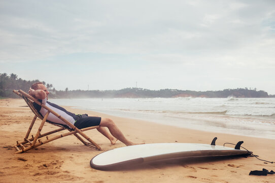 Rainy day underwater case shot of Surfer man sitting on the sandy ocean beach in a comfortable bamboo chair and enjoying the sunset sky. Longboard surf lying in the sand.