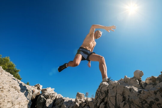 Sky runner with sweaty muscular body running in mountain and jumping over the cleft cliff during the morning jogging. Active sporty people activities wide-angle concept image.