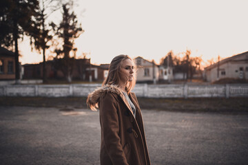 beautiful young blonde model girl. White pants. gray knitted sweater. black boots. . wooden pendant on the neck in the form of a horse. in a brown coat posing. On the Sunset. Portrait. near the trees