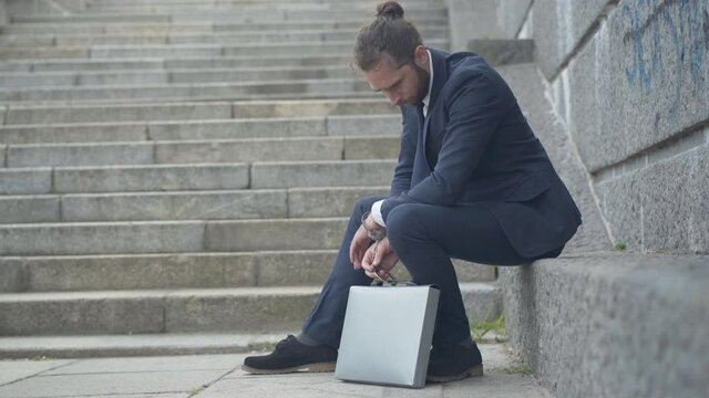 Side view wide shot of confident Caucasian man in handcuffs chained with attache case. Agent sitting on urban city stairs with secret materials. Serious businessman tied to work.