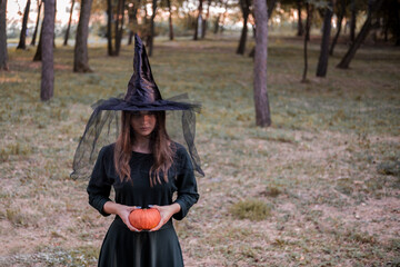Young cute beautiful woman in dark dress and witch's hat holds an orange pumpkin in her hands....