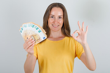 Young woman holding euro banknotes with ok sign in yellow t-shirt and looking happy. front view.