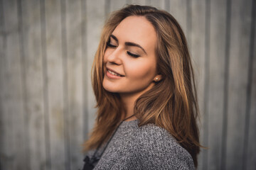 Fototapeta premium beautiful young blonde model girl with eyes closed and smile. gray knitted sweater. On the Sunset. Portrait. best retouch. on the background of gray wooden boards