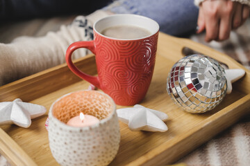 Young woman sits on plaid in cozy knitted woolen white sweater and holds cup of cocoa in her hands. Hygge wooden tray with mug of chocolate, toy tree, candle, stars.