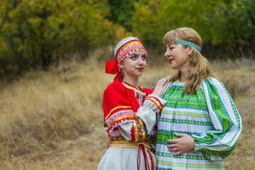 Two young women of different ages in folk costumes of red and green, are hugging each other in the park.