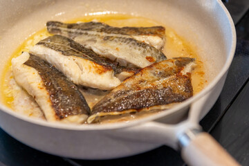 A white frying pan of codfish fillets frying in yellow butter on a stove. The fish fillet has its skin on it, skin side up frying. The portions are all similar in size and have been seasoned with salt