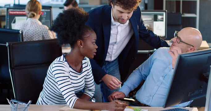 Two office workers: African-American woman and adult European man work in group on common task. Coordinator approaches trainees and gives advice.