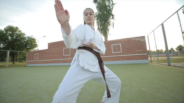 Karate girl in white kimono on the sports ground shows karate techniques and makes a jump with a turn. Close-up. Bottom shooting angle. Slow motion