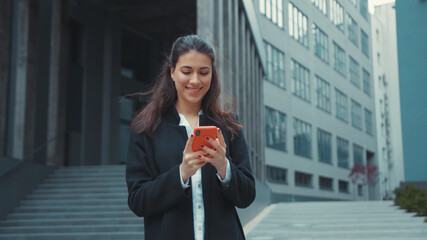 Appealing office young woman walking outside of building using smartphone app networking getting messages on social media smiling cheerful and relaxed.