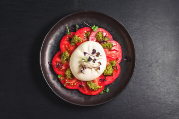 Salad of burrata cheese, tomatoes and pesto sauce in a black plate, on a dark background. Farm food and table setting concept