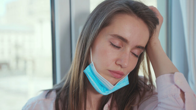 Sad caucasian woman wearing a surgical mask sitting by window at home. Beautiful girl is upset because of coronavirus outbreak. Self-isolation. Lockdown stress.