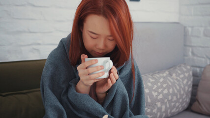 Beautiful sick Asian woman covered in blanket with cute Chihuahua dog resting, warming up by hot drink, recovering at home. Pandemic. Health care. People and pets.