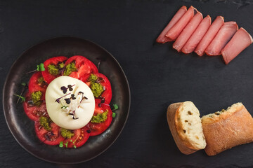 Salad of burrata cheese, tomatoes and pesto sauce in a black plate on a dark surface among meat and bread. Farm food and table setting concept