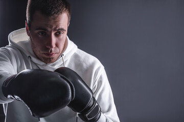 Young man in boxing gloves and a white sweatshirt on a dark background in the studio. The concept of victories and battles