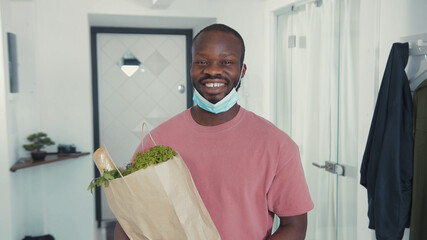 Fototapeta na wymiar Nice african american man wearing protective gloves and face mask, smiling cheerful at camera, holding healthy food package in apartment corridor.