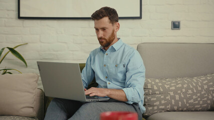 Handsome man using laptop typing working sitting on couch in modern living room at home internet business computer chat smile close up slow motion