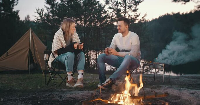 Young man and woman sitting by campfire and drink tea. Traveling hikers male female caucasian couple camping in forest by beautiful lake in evening. Travel together, summer adventure journey in nature
