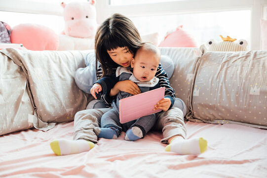 Cute girl and her baby brother sitting on bed and looking at the tablet together
