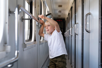 Fototapeta na wymiar Fair-haired boy stands in railway carriage and looks out window. Rail travel
