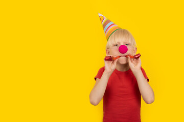 Boy with party hat and red clown nose holding whistle. Portrait on yellow background. Holiday...