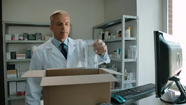 Pharmacist collects order in a box at a pharmacy for online buyer. Caucasian man in white robe preparing goods for delivery. He checking full supervisor medicine list on monitor. 