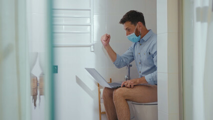 Happy businessman working on laptop wearing mask sit on the toilet at home restroom bearded funny wc isolation quarantine pandemic coronavirus covid-19 portrait close up slow motion