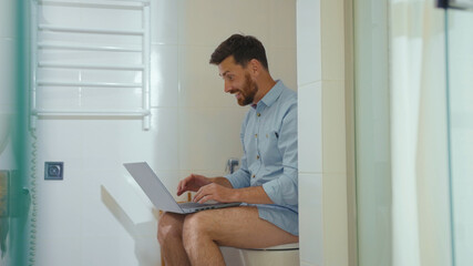 Smiling man workaholic is working in toilet on computer happy reading good news on laptop at home...