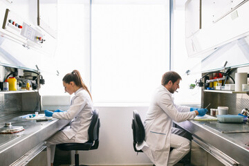 Side view of man and woman in lab coats sitting at tables and examining samples while working in research laboratory together.