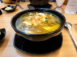 soup served in a black bowl complete with vegetables, seasonings, spoons & forks, asian style food served on a wooden table.