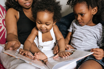 A mother reading with her son & daughter in bed at home. A little boy infant reading out of the book & touching the pages.