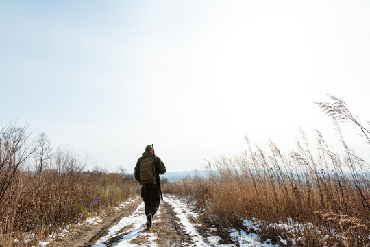 Unrecognizable huntsman walking on snowy countryside road on cold day