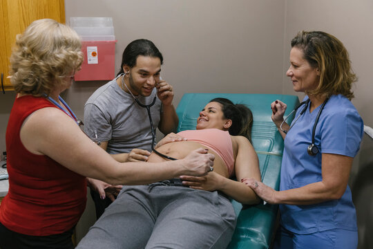 Husband listening to hearbeat of baby in Belly of His wife with Midwife and Doula looking on