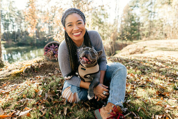 An African American woman sitting down with her french bulldog and playing in the autumn leaves.