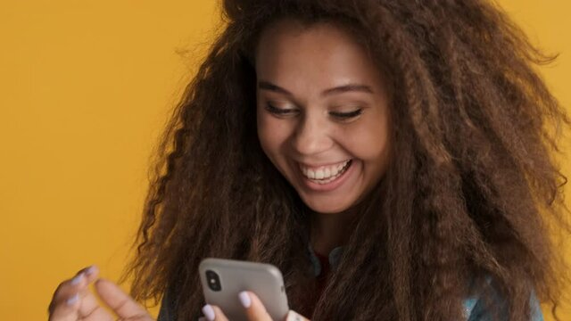 Close up cheerful brunette girl happily chatting online with friends on smartphone being always in touch isolated on yellow background