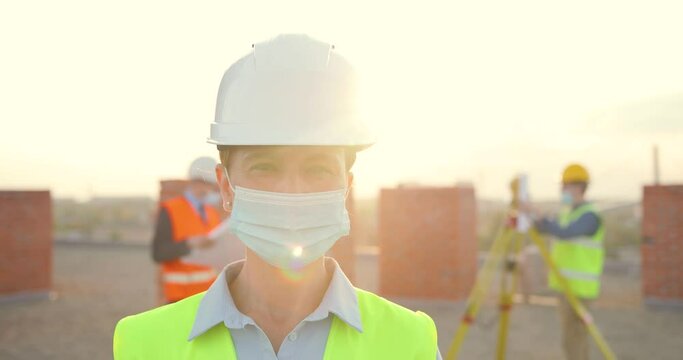 Portrait Caucasian young female constructor in casque and medical mask standing outdoor at construction. Close up of female builder at building in helmet. Coronavirus. Builders working on background.