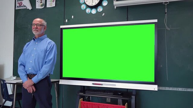 Male teacher stands next to an interactive touchscreen whiteboard with a green screen on it at the front of a classroom set against a blackboard in a school. Remote learning concept.
