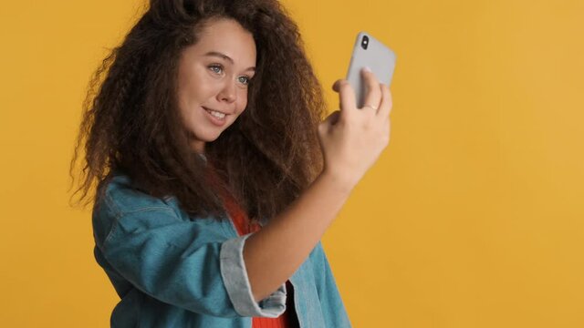 Attractive brunette girl dressed casual wear posing while making selfie on smartphone isolated on yellow background. Modern technology concept 