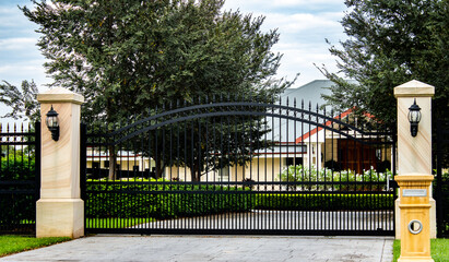 Black metal wrought iron driveway property entrance gates set in brick fence, garden shrubs, trees, lights, house