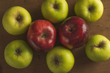 Group of green apples surrounding a couple of red apples on a wooden table.