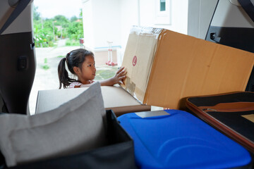 Asian child girl helping parents to carry a cardboard box with stuffs moving into the car to relocation on moving day. Home renovation and relocation concept.