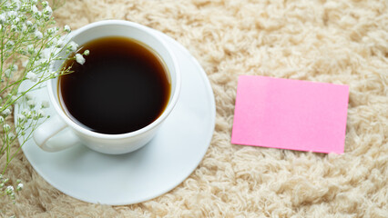 White cup with black coffee placed on a cream carpet. Decorated with white flowers in the corner of the coffee and pink notepaper.