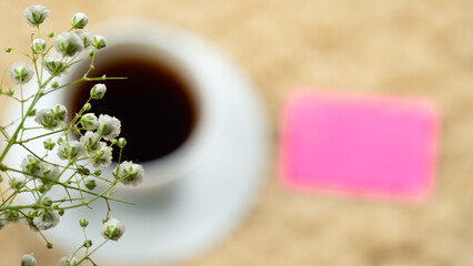 White cup with black coffee placed on a cream carpet. Decorated with white flowers in the corner of the coffee and pink notepaper.
