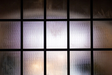 Frosted glass window wall with black metal frame in grid pattern with bright light from the inside.
