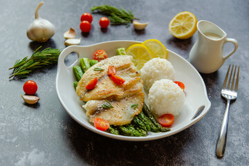 Fried tilapia, asparagus, cherry tomatoes, lemon and rice balls on a white plate.