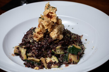 Traditional Spanish seafood chicken meat greens paella with zucchini and cheese on a dark wooden table. Fried shrimps, squids. Vegan, vegetarian. Black brown rice. Healthy food on a white plate.