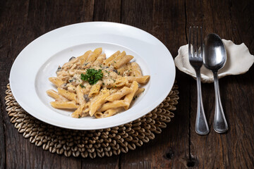 Homemade delicious pasta macaroni with white mushrooms chicken meet mushrooms in creamy cheese sauce in the white plate on the dark brown wood table. Parsley, spicy. Yellow, green.