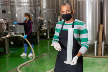 Portrait of male winery worker in mask controlling winemaking process, checking wine near metal tanks for wine fermentation